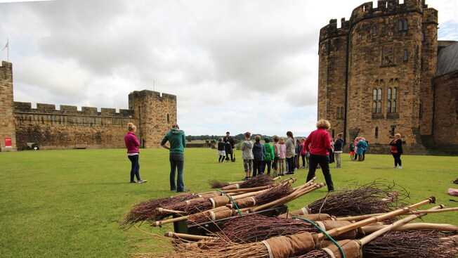 A pile of broomsticks made of branches next to a man coaching kids on a castle field