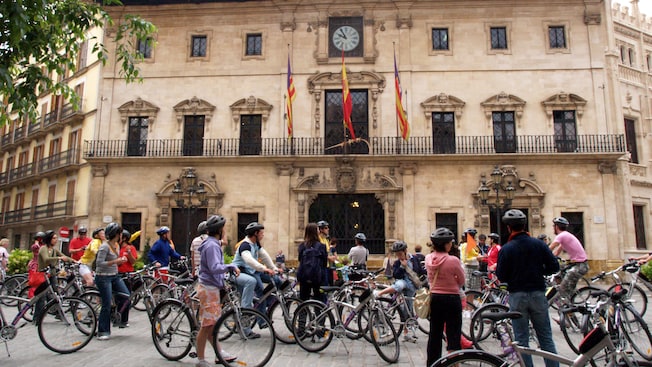 People standing by bicycles in front of a building featuring flags, a clock, windows and more People standing by bicycles in front of a building featuring flags, a clock, windows and more