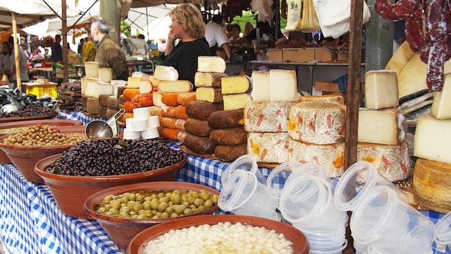 A street market featuring a kiosk with hanging sausage, rounds of various types of cheese and terracotta bowls filled with olives