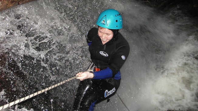 A woman, dressed in a wetsuit and helmet, rappels into a torrent of water