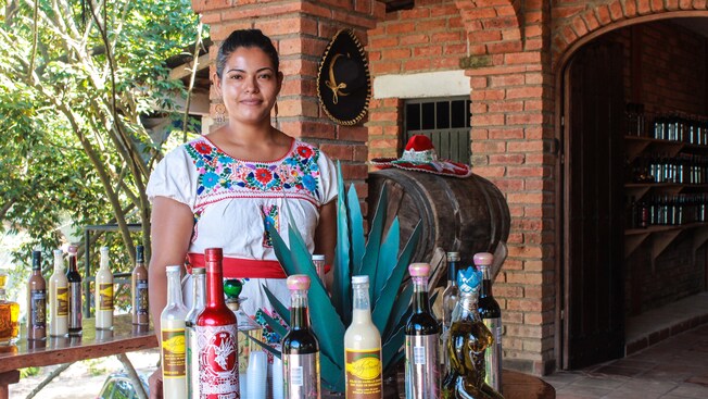 A young woman dressed in traditional Mexican attire smiles while standing next to a selection of tequilas