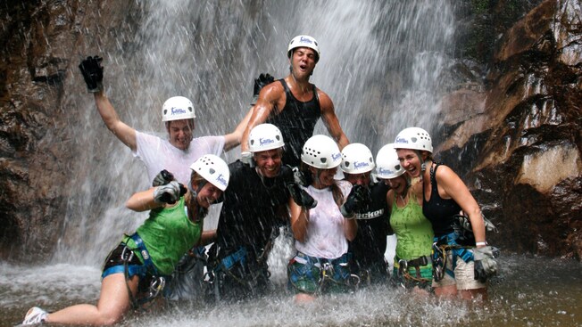 Excited men and women, wearing helmets, pose for a picture under a waterfall