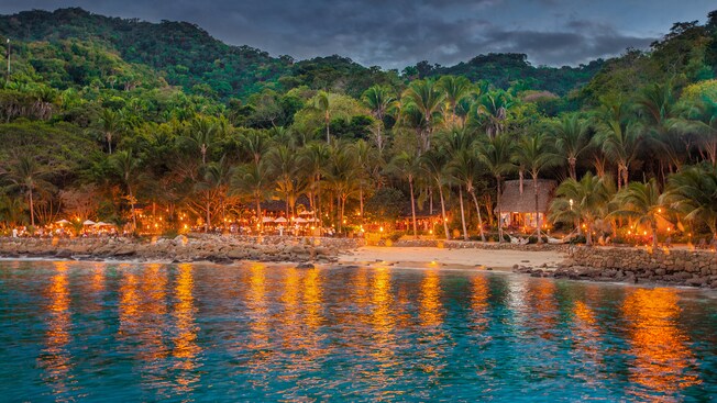 A beach cove with an outdoor restaurant is surrounded by lush mountains and palm trees