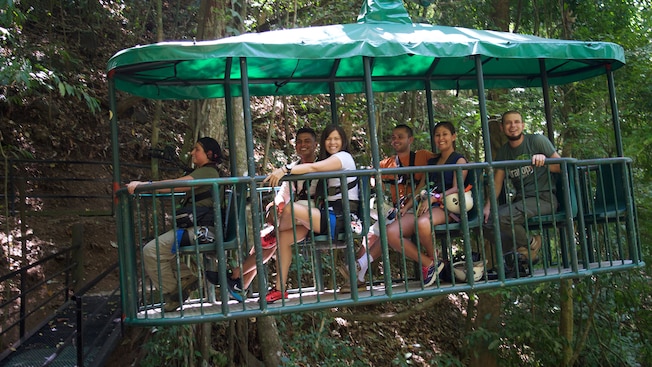 A tour guide and 5 guests smile while sitting on board an open air gondola in the rainforest A tour guide and 5 guests smile while sitting on board an open air gondola in the rainforest