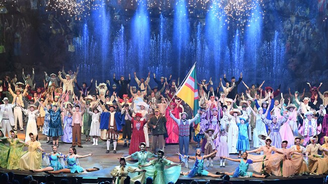 A group of performers dressed in 19th century attire on a stage with fireworks and fountains of water