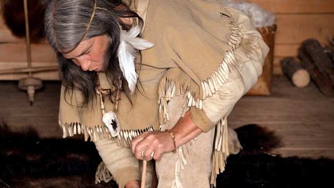A Canadian Indian wearing traditional clothes leans over holding a stick to the ground