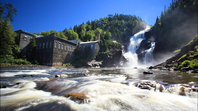 A waterfall falls between 2 hills and spills into a pool of raging water near a building