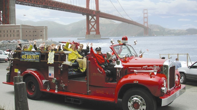 People wave from every seat of a vintage, roofless fire engine with the Golden Gate Bridge in the background