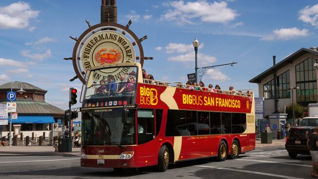 People ride in a red double decker bus with no roof on its second floor in front of a Fishermans Wharf sign