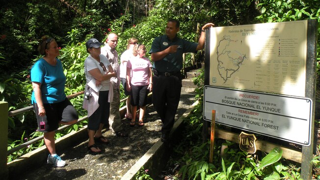 A tour group leans against a railing in a forest while a forest ranger points to a map board