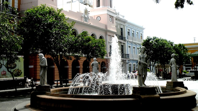 A fountain with 4 statues placed evenly around the rim sits in a plaza lined by trees A fountain with 4 statues placed evenly around the rim sits in a plaza lined by trees