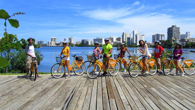 Un grupo de adultos y niños con bicicletas sonríe a la cámara en un paseo marítimo de madera