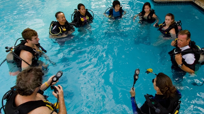 A group of scuba students stand in a swimming pool and gather around a dive instructor while learning how to use their equipment