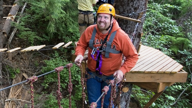 A man outfitted in a helmet and harness smiles as he walks along a suspended bridge in the tree tops A man outfitted in a helmet and harness smiles as he walks along a suspended bridge in the tree tops