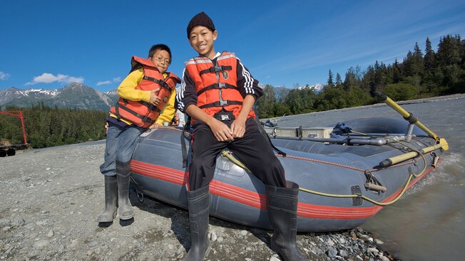 Two young boys in life vests pose with an inflatable raft on the rocky shores of a mountain river