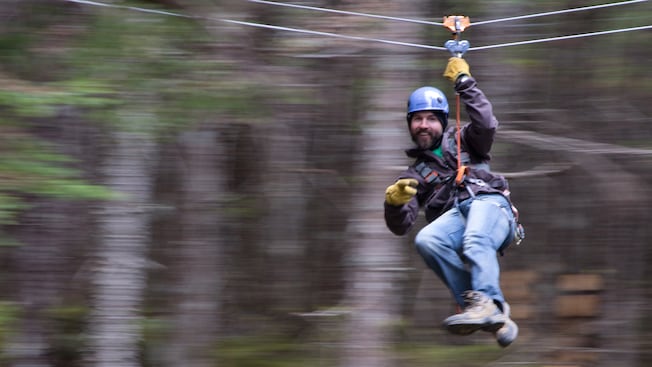 Um homem com barba usando capacete, arnês e luvas sorri enquanto passa de tirolesa pela floresta Um homem com barba usando capacete, arnês e luvas sorri enquanto passa de tirolesa pela floresta