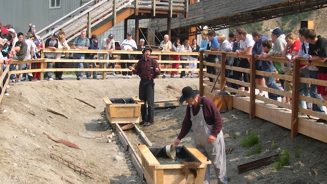 A tour group stands around a shallow dirt pit watching a re-enactment of 2 prospectors panning for gold