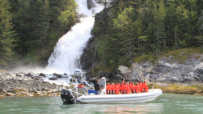 A tour group donned in waterproof suits watch a rushing waterfall as they sit in a stopped rigid inflatable boat
