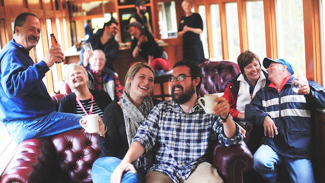 A tour group laughing and smiling while sitting in overstuffed chairs and holding up a variety of beverages inside a rail car A tour group laughing and smiling while sitting in overstuffed chairs and holding up a variety of beverages inside a rail car