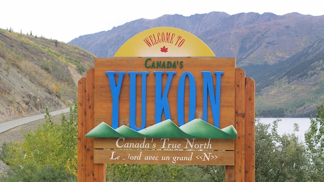 A wooden sign alongside a mountain road and river reads "Welcome to Canada's Yukon--Canada's True North" A wooden sign alongside a mountain road and river reads "Welcome to Canada's Yukon--Canada's True North"