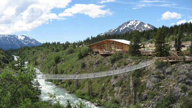 A suspended pedestrian bridge over a rushing river leads to a large wooden visitors center