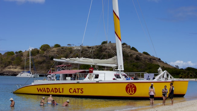 Um grupo de pessoas caminha na parte rasa de uma praia enquanto um grande catamarã está parado na água atrás deles