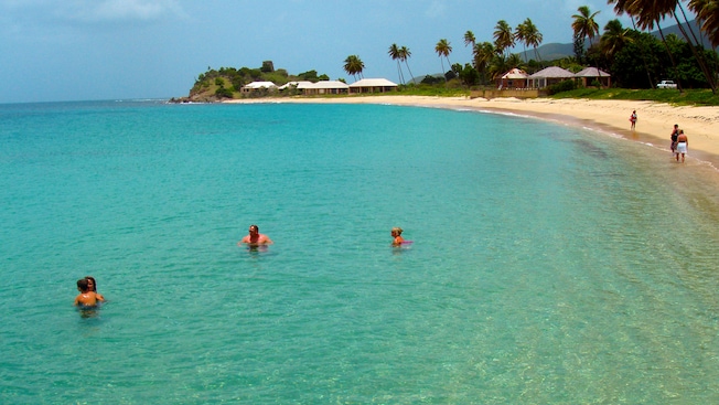Tres personas en el mar, con el agua hasta el pecho, cerca de una playa rodeada de palmeras y pequeñas edificaciones Tres personas en el mar, con el agua hasta el pecho, cerca de una playa rodeada de palmeras y pequeñas edificaciones