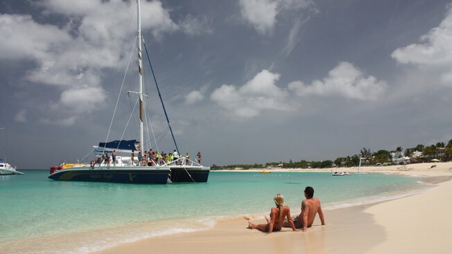 Una pareja se recuesta en la arena de una playa tropical, mientras que un catamarán se encuentra anclado cerca de ellos