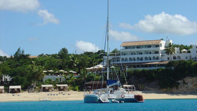 A catamaran docked close to a shoreline of a tropical beach with cabanas, a restaurant and a hotel