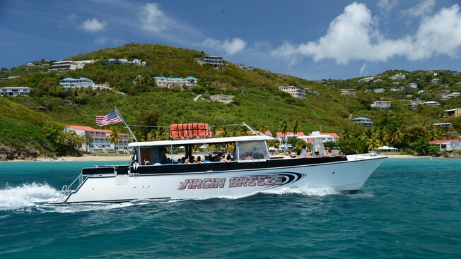A boat called the Virgin Breeze sails past a tall resort building near the shore