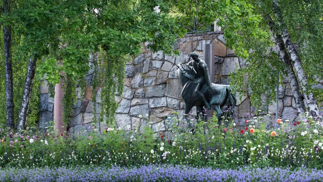 A statue of a man on a horse near flowers, trees and a wall
