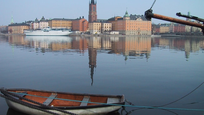 City buildings line the shore and across a wide river, a row boat is tied to a dock