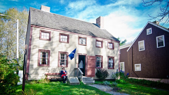 A man wearing clothing reminiscent of American colonists sits outside a house of the period