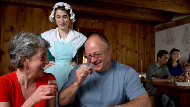 A woman dressed in a colonial costume smiles while watching a man and a woman hold shot glasses