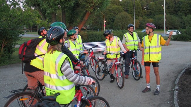 A group of people wearing safety vests and helmets stand with their bicycles listening to a guide