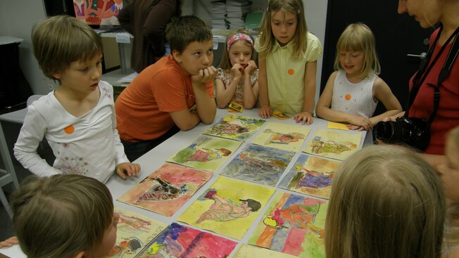 A woman talks to kids who are gathered around colorful illustrations on a table
