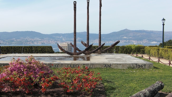Stone platform with rectangular fountain and a sculpture of 3 anchors overlook a bay and coast line