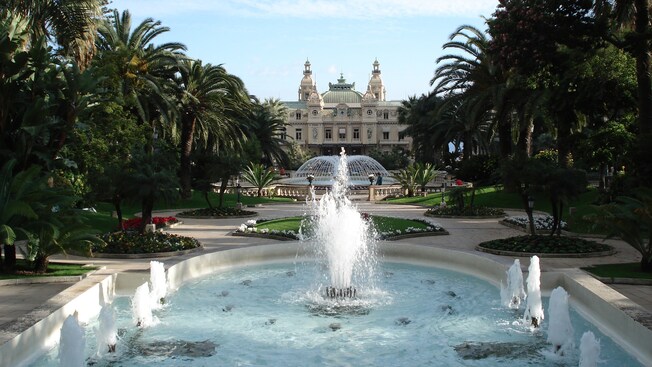 Um edifício grande e ornamentado tem vista para uma grande fonte circular, várias plantadeiras e duas fontes