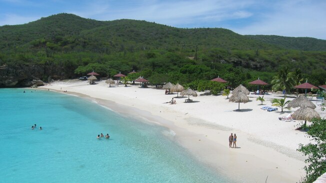 A gorgeous sandy beach is dotted with umbrellas, palm trees and vacationers with lush mountains nearby