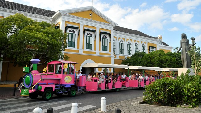 A passenger tram drives by one of Willemstads Dutch colonial buildings surrounded by lush foliage