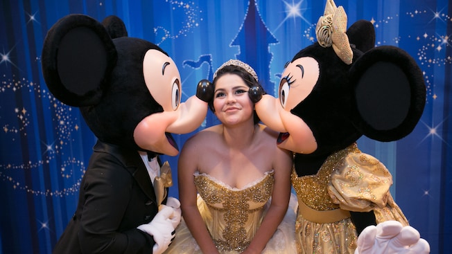 A smiling young woman in a quinceñera gown and tiara bends to let Mickey and Minnie Mouse plant a kiss on each of her cheeks