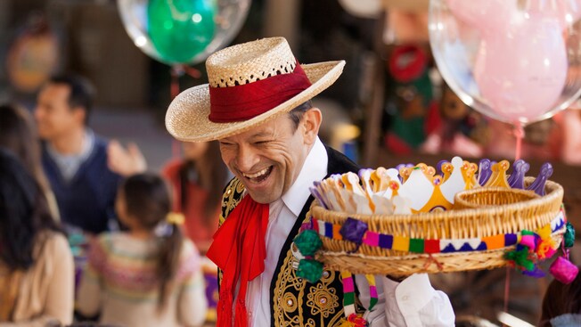 At a 3 Kings Day celebration, a man in festive Latin American dress carries a tray containing paper crowns