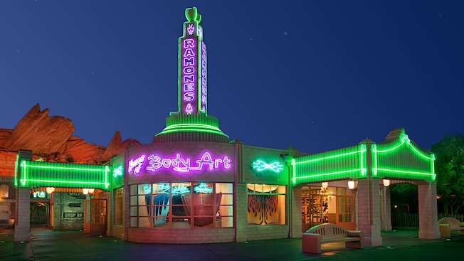 A shop that resembles a vintage gas station lit up at night by neon lights with signs that read "Ramones" and "House of Body Art" and featuring windows displaying custom-designed automobile hoods