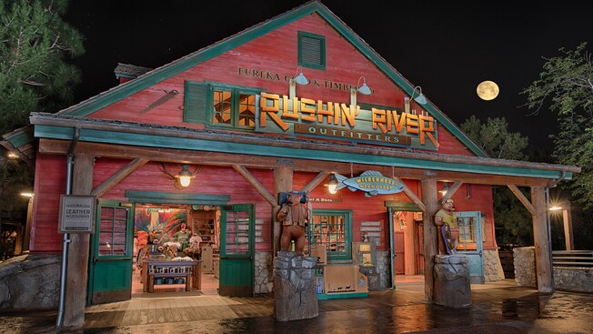 A rustic shop surrounded by trees with carved wooden bears on the porch and signs that read 'Rushin' River Outfitters', 'Eureka Gold & Timber Co.' and 'Wilderness Wear & Gear', selling apparel and hats themed to California and Grizzly River Run