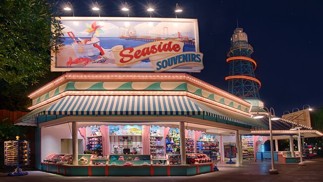 An open-air shop selling merchandise that includes sunglasses, Mickey Mouse ear hats, novelty gloves, pins, toys and snacks, beneath a lighthouse-like tower and a huge billboard sign with an illustration of a crab, a bucket of seashells, a beach, an ocean pier and the words "Seaside Souvenirs"
