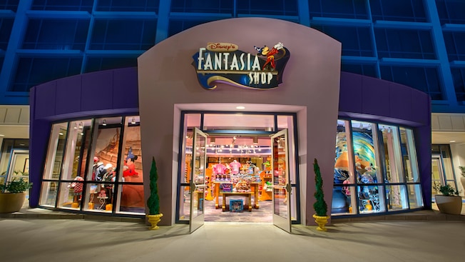 A shop at the base of a modern hotel tower with a sign featuring Mickey Mouse that reads 'Disney's Fantasia Shop' and merchandise that includes Disney-themed apparel, dolls and toys