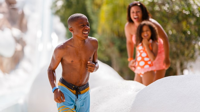 A smiling boy pops up from the waterslide while his mother and younger sister look on