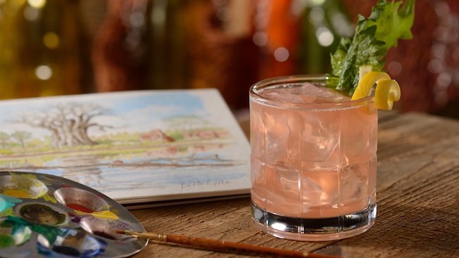 An icy glass with a Spice Trader cocktail garnished with a mint leaf and lemon peel curl on a table beside a painters pallet and watercolor painting of an Asian forest scene