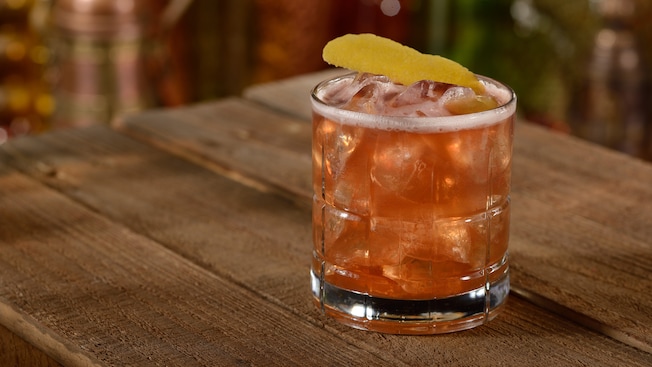 An icy glass with a Tempting Tigress cocktail garnished with a slice of lemon peel on a wooden table