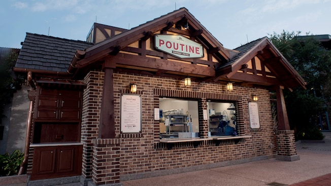 The front of the Daily Poutine restaurant in Disney Springs features a brick façade with 2 serving windows, a sign bearing the establishment’s name and a twin-peaked roof